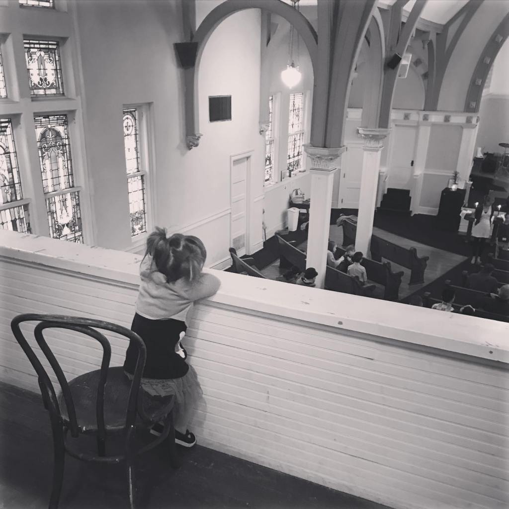 A grayscale photo of a young girl sitting on a wooden chair in the balcony of a church, peering over the edge to watch the Easter worship service below. The church has stained glass windows and wooden pews.
